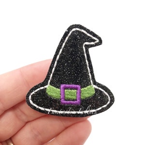 May include: A black glitter witch hat patch with a white stitched outline. The hat features a green band with a purple buckle. The text "Fantastic Felties" is visible on the bottom.