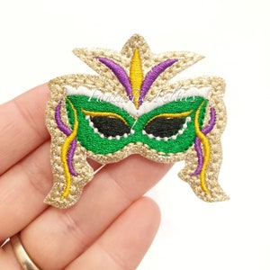 May include: Green and gold Mardi Gras mask with purple accents. The mask is embroidered with glitter and has a feather design on top.