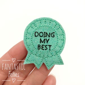 May include: A green felt award ribbon with a white glitter finish and black embroidered text that reads "Doing My Best".