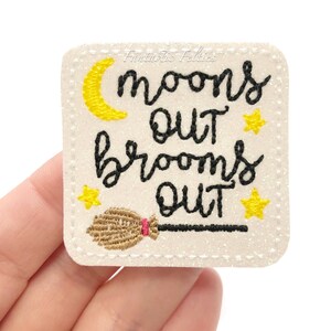 May include: A square, off-white felt patch with the text "moons out, brooms out" in black script. A yellow crescent moon and stars accent the text. A brown broom with a pink handle is at the bottom. The patch is edged with white stitching.