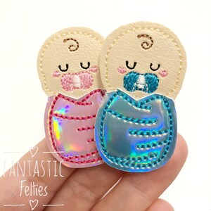 May include: Two felt baby appliques, one in pink and one in blue, with embroidered faces and pacifiers. The appliques are made of iridescent fabric.