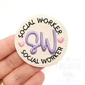 May include: White felt circle with black stitching around the edge. The text "Social Worker" is written around the circle in black. The letters "SW" are embroidered in purple in the center of the circle. Two pink hearts are embroidered on either side of the "SW".