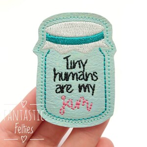 May include: A light blue felt patch shaped like a jar with the text "Tiny humans are my jam" in black and pink embroidery.