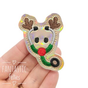May include: A reindeer-themed applique with a stethoscope design. The applique features brown antlers, a red nose, and a green and silver stethoscope. The text "Fantastic Felties" is visible in the lower left corner. The applique is held in a hand.