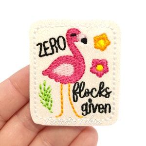 May include: A square, off-white felt patch with a pink flamingo design and the text "ZERO flocks given." The flamingo has a black beak and orange legs. There are also embroidered flowers and greenery.
