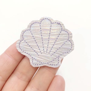 May include: A white iridescent seashell with a purple stitched outline. The seashell is made of faux leather and has a textured surface.
