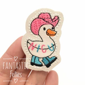 May include: An embroidered patch featuring a white duck wearing a pink cowboy hat and blue boots. The duck has an orange beak and the word "NICE" in pink and blue across its chest. The patch is on a white background with the text "FANTASTIC Felties".