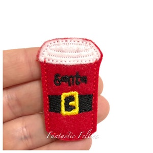 May include: A red felt Christmas-themed coffee cup with the word "Santa" in black script. The cup features a black belt with a yellow buckle and a white foam top. The words "Fantastic Felties" are at the bottom.
