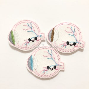May include: Three white felt eye patches with pink stitching and embroidered details. Each eye patch has a different colored iris: green, brown, and blue. The eyes have black eyelashes and a smiling mouth.