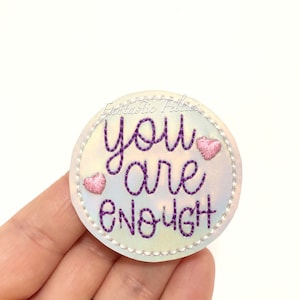 May include: A circular patch with a pearlescent white background featuring the embroidered purple text "you are enough" in a cursive font. Two small pink embroidered hearts are placed on the left and right sides of the text.