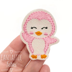 May include: A pink and white felt penguin with a glittery white body and pink wings. The penguin has a yellow beak and orange feet.