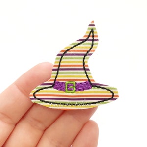May include: A colorful witch hat with horizontal stripes in orange, yellow, green, purple, and white. The hat features a purple belt with a green buckle and black stitching. The text "Fantastic Felties" is printed on the bottom.