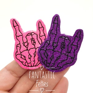 May include: Two felt skeleton hand patches in a rock and roll hand gesture. The patches are pink and purple with black glitter.
