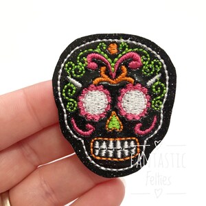 May include: A black embroidered skull patch with white, pink, green, and orange details. The patch has a glittery finish and a white stitched border. The design features floral and decorative elements, perfect for Halloween or Day of the Dead.