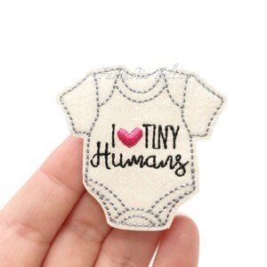 May include: White glitter iron-on patch with a gray outline of a baby onesie. The onesie has a pink heart and the text "I ♥ TINY HUMANS" in black embroidery.