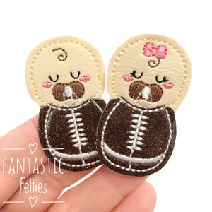 May include: Two felt baby appliques in the shape of footballs. The appliques are brown with white stitching and have a baby face with closed eyes and a pacifier in their mouth. One applique has a pink bow on top of its head.