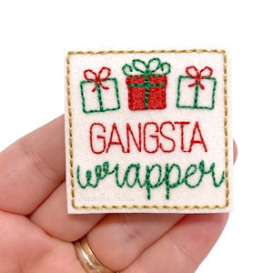 May include: A white felt patch with gold stitching around the edges. The patch features three red and green embroidered Christmas presents and the text "Gangsta wrapper" in red and green thread.