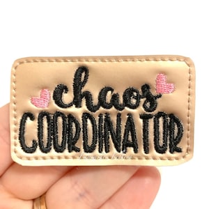 May include: A gold-colored fabric patch with black embroidered text that reads "chaos coordinator" and two pink embroidered hearts.
