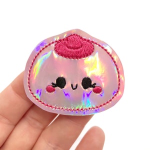 May include: A pink iridescent iron-on patch with a cute cartoon face and a pink embroidered top knot. The patch says "Fantastic Felties" at the bottom.