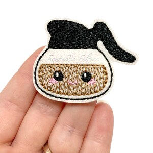 May include: A white felt applique of a coffee pot with a black lid and brown embroidered details. The coffee pot has a pink blush and black eyes. The text "Fantastic Felties" is visible in the background.