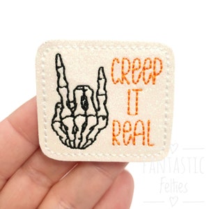 May include: A square, off-white patch with a black skeleton hand making a rock-and-roll gesture. The phrase "CREEP IT REAL" is embroidered in orange thread. The patch has a glittery texture and a stitched border.