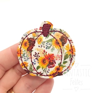 May include: A white felt pumpkin with a floral pattern featuring sunflowers, burgundy roses, and green leaves. The pumpkin has a burgundy felt stem and is stitched with a brown thread.