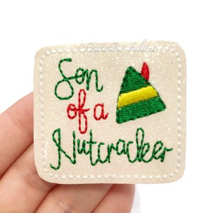 May include: White felt patch with green glitter. The patch has red and green embroidery that says "Son of a Nutcracker" with a green elf hat with a yellow band and red tip.