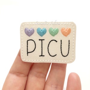 May include: A rectangular, off-white felt patch with the word "PICU" embroidered in black. Above the text are four embroidered hearts in pastel colors: purple, teal, green, and orange. The patch has a glittery finish and a stitched border.