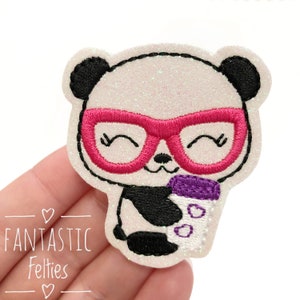 May include: A white panda with black ears and eyes wearing pink glasses and holding a purple and white cup with hearts on it. The panda is embroidered on a white glittery fabric.