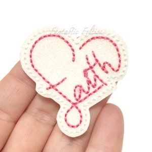 May include: A white felt heart with pink stitching. The word "Faith" is embroidered inside the heart.