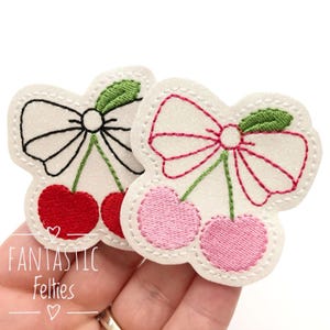 May include: Two felt patches with embroidered cherry designs. One patch features a black bow and red cherries, while the other has a pink bow and pink cherries.