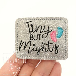 May include: A silver glitter iron-on patch with the text "Tiny but Mighty" in black cursive font. Two embroidered footprints, one pink and one blue, are to the right of the text.