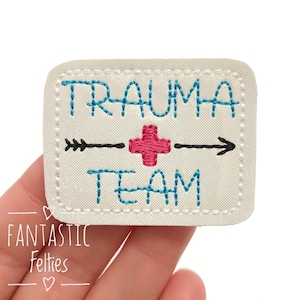 May include: White felt patch with blue embroidery that reads "Trauma Team" with a red plus sign and black arrow in between the words.
