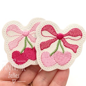 May include: Two felt cherry patches with pink and red bows. The cherries are pink and red and have green stems. The patches are white with a glittery texture.