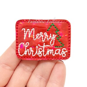 May include: A red rectangular Christmas-themed patch with the words "Merry Christmas" embroidered in white thread. A green embroidered Christmas tree is on the right side. The patch is held in a hand.