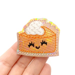 May include: A sparkly orange and white embroidered patch of a smiling pumpkin pie slice with a white whipped cream topping. "Fantastic Fables" is written in white text at the bottom of the patch.