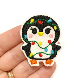 May include: A felt penguin patch with black, white, orange, pink, and green embroidery. The penguin is decorated with a string of colorful Christmas lights. The patch is a festive holiday decoration.