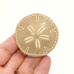 May include: A beige felt sand dollar with a white embroidered star design. The sand dollar is covered in glitter.