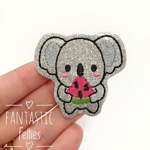 May include: A silver glitter koala holding a slice of watermelon. The koala is embroidered with black outlines and pink cheeks. The watermelon is embroidered with green, red, and black.