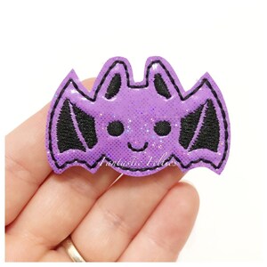 May include: A purple bat-shaped felt applique with black embroidered details. The bat has a smiling face and glitter accents. The text "Fantastic Felties" is visible on the bottom. This item is suitable for crafting and embellishment.