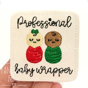 May include: White felt patch with embroidered text "Professional baby wrapper" and two cartoon figures, one in red and one in green.