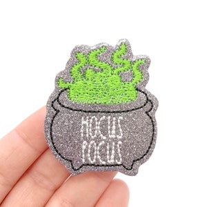 May include: A glittery gray cauldron patch with green flames and the words "Hocus Pocus" embroidered in white. The patch is a Halloween-themed accessory, perfect for adding to clothing or bags.