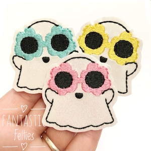 May include: Three white felt ghost appliques with embroidered black eyes and mouths. Each ghost is wearing a pair of sunglasses made of felt flowers in different colors: turquoise, yellow, and pink.