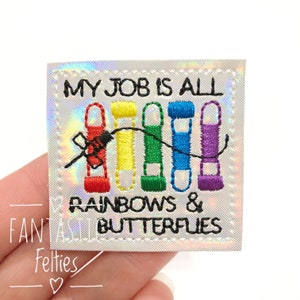 May include: Embroidered patch with the text "MY JOB IS ALL RAINBOWS & BUTTERFLIES." The patch features colorful spools of thread in red, yellow, green, blue, and purple, connected by a black thread. The patch has a holographic background.