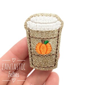 May include: A gold glitter coffee cup with a white lid and a small orange pumpkin with a green stem on the front. The cup is embroidered with brown stitching.