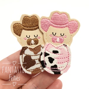 May include: Two felt cowgirl finger puppets, one with a brown cowboy hat and brown and white cow print, the other with a pink cowboy hat and pink and white cow print.