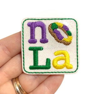 May include: A white felt patch with green stitching around the edges. The patch features the letters "NOLA" in purple, green, and yellow embroidery. A purple, green, and yellow embroidered donut is between the "N" and "O".