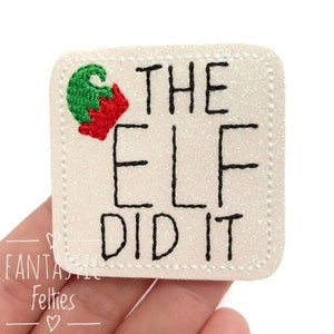 May include: A square, glittery patch with the text "THE ELF DID IT" in black, stitched lettering. A small embroidered elf hat in green and red is in the upper left corner. The patch is off-white with a dotted border.