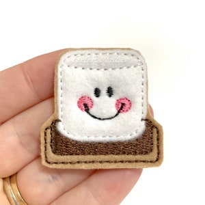 May include: A felt applique of a smiling marshmallow with pink cheeks, on a brown felt base, resembling a s'more. The text "Fantastic Felties" is visible in the top right corner.