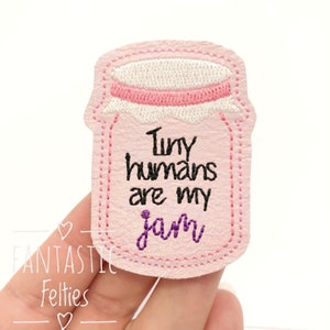 May include: Pink felt applique shaped like a jar with the text "Tiny humans are my jam" in black and purple embroidery.